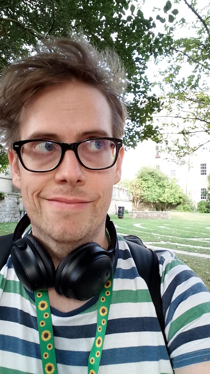 An image of a 36-year old white autistic gayboy, wearing glasses, stubble, headphones around his neck, and the bands of his rucksack over a stripy t-shirt.