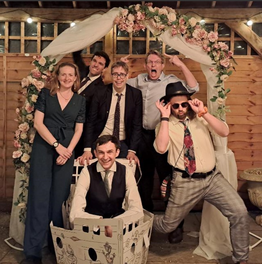 An image from a wedding reception. Five people in their thirties are positioned underneath a gazebo. The bridegroom (white, dark hair, dressed up to the nines) is in a cardboard boat, in the centre. With him, a thirty-something year old white woman smiles kindly. Next to her, a thirty-something year old dark-haired white man in a suit is biting his lower lip as he appears to pull down the gazebo. In the centre of the image is Jorik, a 36-year old autistic white gay in a suit wearing his father-in-law's tie, with a face that describes a self-insert teacher character in an Alan Bennett play. To my other side, a white man wearing glasses giving double middle fingers and wearing a facial expression of deep rage. The last man in his thirties is wearing a hat, dark sunglasses and is leaning in a fashion reminiscent of a human doing an impression of a motorbike. These were my colleagues when I was teaching English as a Foreign Language in Oxford, 2017-2020.