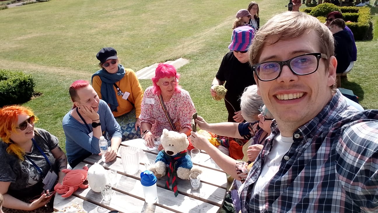 A picture of a white autistic man in his mid-to-late thirties, wearing glasses and a stripy shirt, from an unflattering angle. The other 7 people are sitting at a picnic table with freshly mown grass behind them. Everyone is autistic, from a variety of backgrounds and colours of hair.