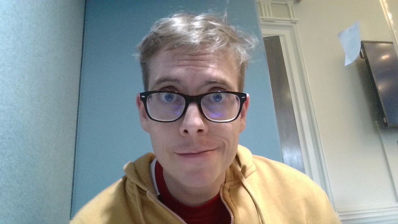 A snapshot of a pale-looking 36 year old white autistic gay man, wearing black-rimmed glasses and a yellow hoodie. The man's eyebrows are raised, his mouth closed in a diagonal-esque shape.