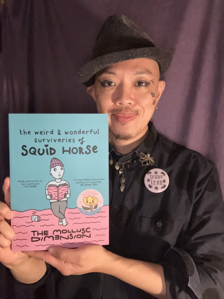 An image of Squid Horse AKA The Mollusc Dimension. Squid is a neurodivergent human from the East Asian diaspora, with a moustache and slight beard on his chin. He has a shaved head and wears a bowler hat askance, folding his right hear. He wears eyeshadow and eyeliner with crow's feet that extend, as well as painted coloured stars. He is wearing a black shirt, with a necklace, a brooch and a button saying 'trans icon' in a rune-like font. He is looking directly into the camera and holds his book 'the weird & wonderful surviveries of Squid Horse' aloft, smiling as he does so.