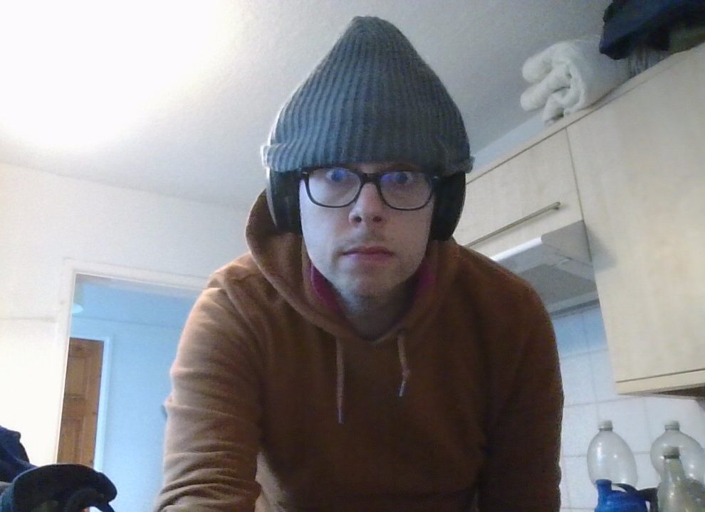 An image of Jorik, a 37-year old autistic gay man in his kitchen in January 2025. Jorik wears a comfy winter hat over black glasses and headphones, as well as a cocoa-coloured hoodie.