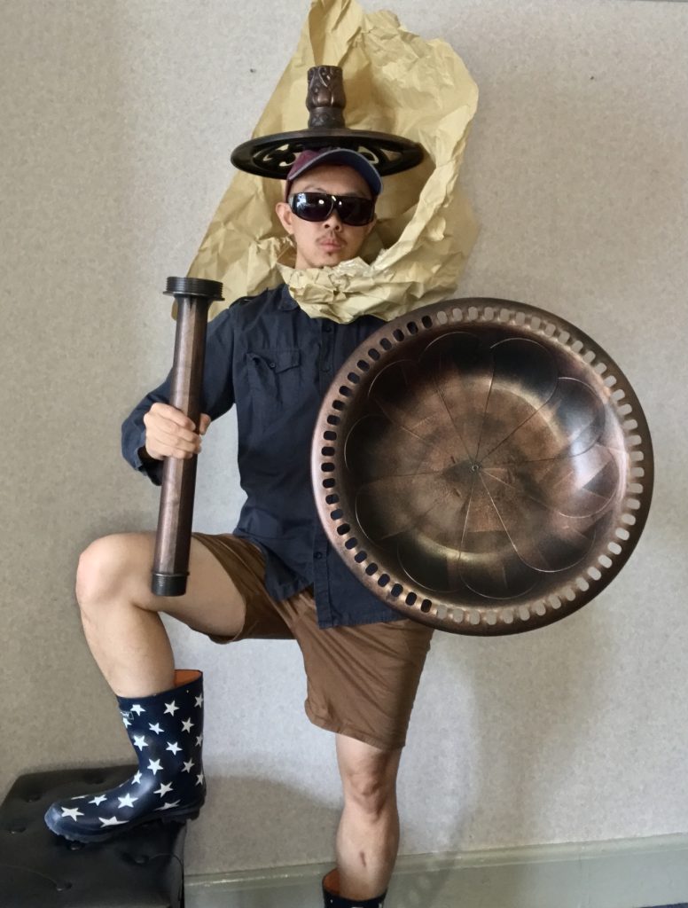 An image of a neurodivergent British East-Asian diasporic trans masculine person in his thirties, wearing what can only be described as takeaway gladiator outfit, with star-coverd wellies, a dark blue navy shirt, brown shorts, a light fitting for a weapon, something odd and bronze as a shield, huge sunglasses, a baseball cap and something that looks like an inverted bronze ceiling rose with yellow/beige coloured paper exploding out of it like the Trevi fountain, encircling Squid's face. Not asking why.